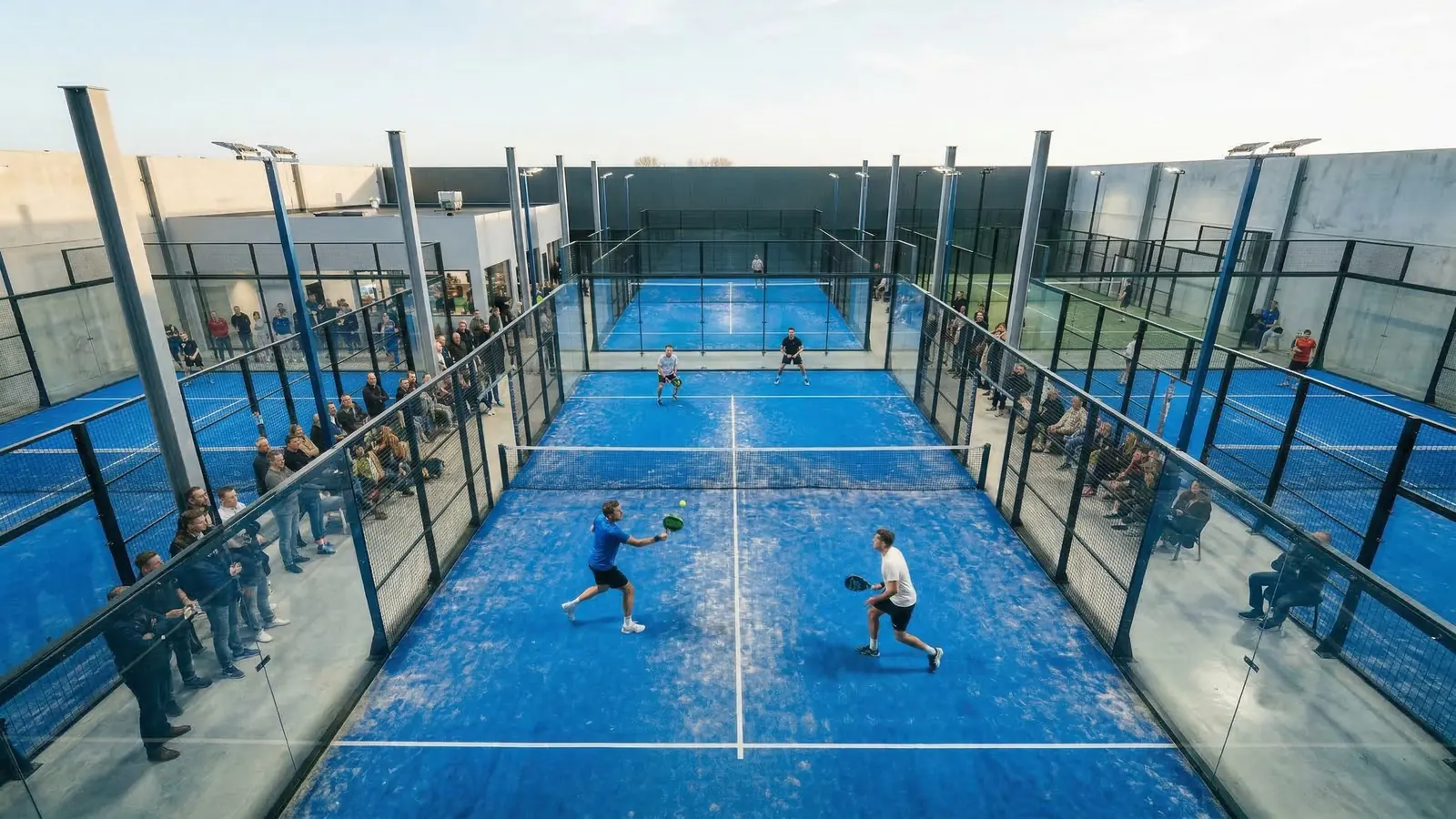 Deux paires s'affrontent sur un court de padel vitré, entourées de spectateurs